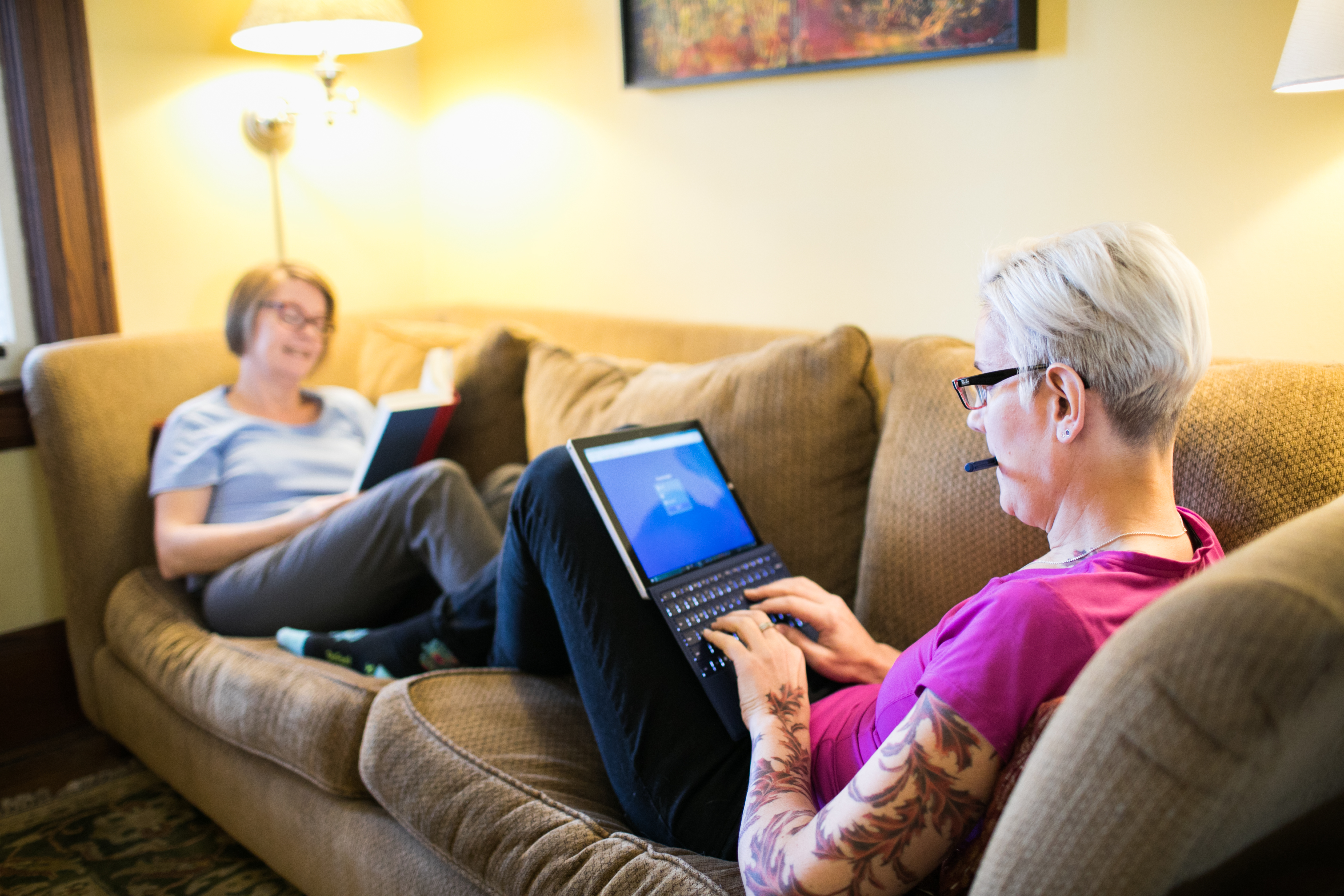 Two women sit on the couch while one reads a book and the other works on a laptop computer