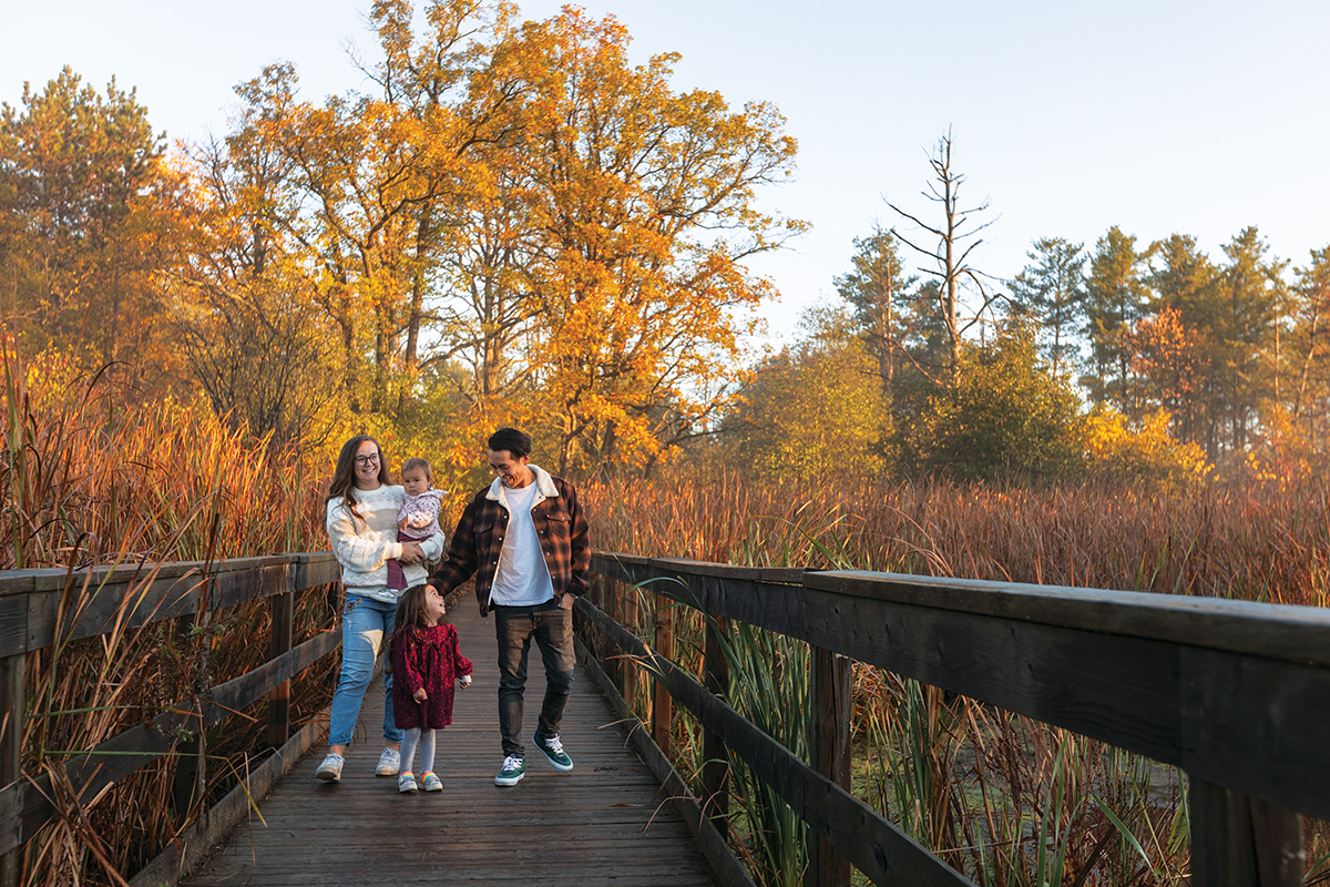 Photo d’une famille marchant sur un trottoir de bois, entouré d’herbes hautes et d’arbres au feuillage coloré en automne.